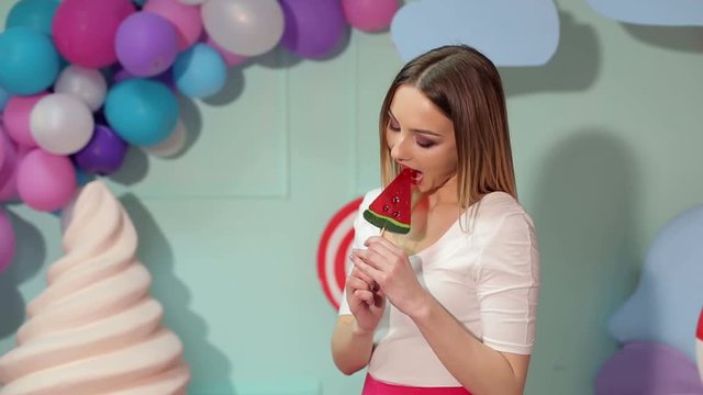 Portrait of a happy girl with a big candy on a background of multi-colored balls, she wants to bite the candy and can't decide. Diet. Barbie. Lollipop.