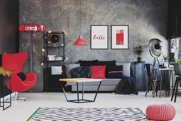Red lamp above table in front of sofa in dark living room interior with posters and armchair. Real...