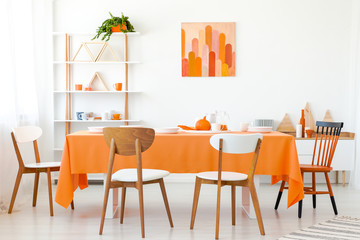 Wooden chairs at orange table in white dining room interior with poster and plant. Real photo