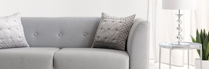 Metal end table with glass lamp placed by the light grey lounge with two silver cushions in real photo of white living room interior