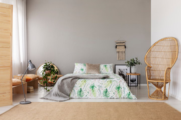 Wicker peacock chair in stylish bedroom interior with natural carpet and king size bed, real photo with copy space and macrame on the empty grey wall