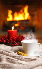 Cup of hot drink with steam and cookie berries red candle in Christmas decoration on knitted plaid in front of fireplace. Christmas New Year concept. Cozy relaxed magical atmosphere home interior.