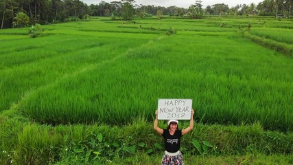 Happy New Year 2019. 4K flying drone video of young woman with santa hat and whiteboard with handwritten text posing among green rice field on Bali island. - Powered by Adobe