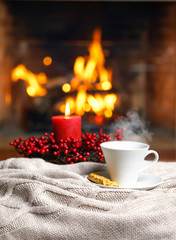 Obraz na płótnie Canvas Cup of hot drink with steam and cookie berries red candle in Christmas decoration on knitted plaid in front of fireplace. Christmas New Year concept. Cozy relaxed magical atmosphere home interior.