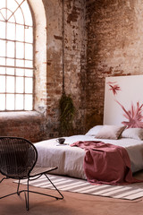 Armchair in front of bed with red blanket in industrial bedroom interior with painting and window. Real photo