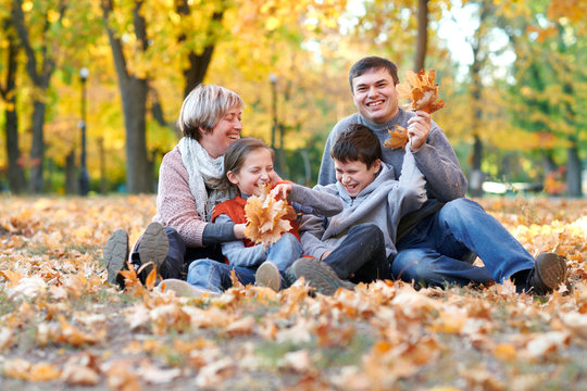 Happy family sit in autumn city park on fallen leaves. Children and parents posing, smiling, playing and having fun. Bright yellow trees.