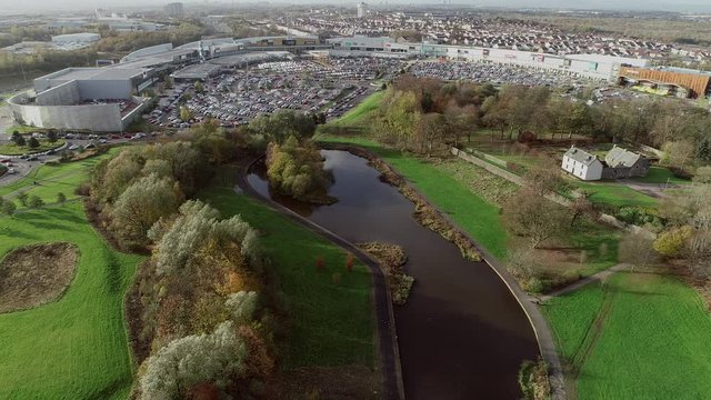 Aerial footage over the pond in Auchinlea Park in the East of Glasgow to Provan Hall, a medieval fortified country house, surrounded by trees, and the Glasgow Fort retail and leisure park. 