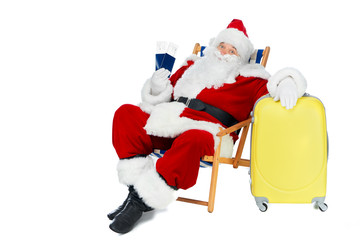 santa claus holding two passports and air tickets for christmas trip while sitting in beach chair with travel bag isolated on white