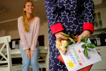 Child congratulates mother on Mother's Day, hides behind a gift with a postcard and flowers
