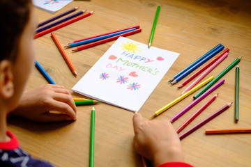 A boy sits at a wooden table on which is a Mother's Day postcard and colored pencils