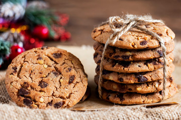 Chocolate chip cookies freshly baked are stacked on sackcloth with fir and gift festive decoration on wooden table background. Christmas cookies. Homemade pastry.