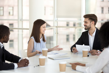 Millennial workers sit at table in boardroom having friendly talk during briefing, smiling young employees laugh negotiating or brainstorming in office, coworkers have casual discussion at meeting
