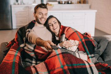 happy young couple watching tv with popcorn on couch and covering with plaid - Powered by Adobe