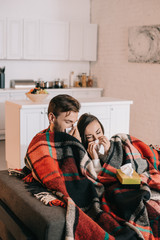 diseased young couple with paper napkins sneezing while sitting on couch under plaid at home