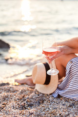 Girl is sitting on sea beach with wineglass of wine at sunset in summer vacation in resort. Tourist...