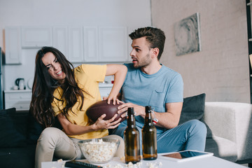 angry young woman fighting for american football ball with husband during match on tv at home