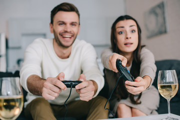 emotional young couple with glasses of wine playing retro video game on couch at home