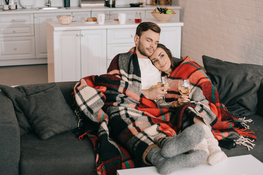beautiful young couple with glasses of white wine relaxing on couch under plaid