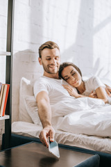 happy young man relaxing in bed with girlfriend and reaching for smartphone