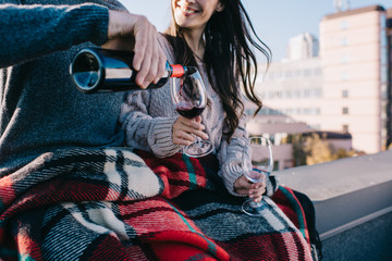 cropped shot of young couple covering in plaid on rooftop and pouting red wine into glasses