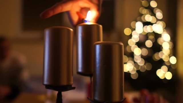 Male hand lights the candles. Close up of three candles with blurred Christmas light background