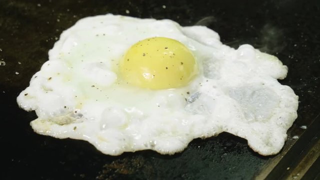 Close-up shot of broken egg being salted and peppered for frying on hot oiled grill. 4K