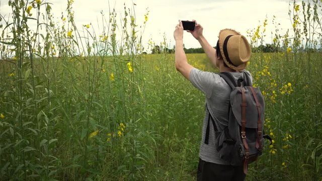 Young Asian tourist backpacker wearing hat and carrying a bag taking photos on the countryside glassland during holiday vacation