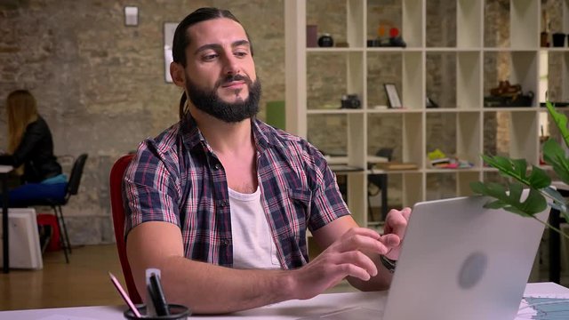 Footage of calm and happy nice caucasian guy in casual wearing with dark beard who is sitting near his laptop and looking at camera chill after a while, working vibes, indoor