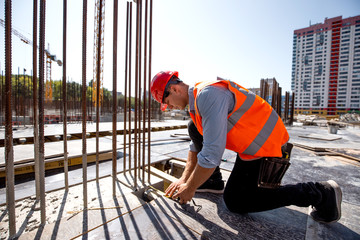 Civil engineer dressed in shirt, orange work vest and helmet measures the hole with a tape measure on the building site