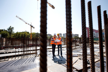 Two men dressed in shirts, orange work vests and helmets explore construction documentation on the...