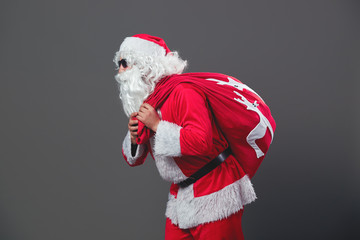 Santa Claus with sunglasses runs with the bag in Christmas gifts on his back on the white background.