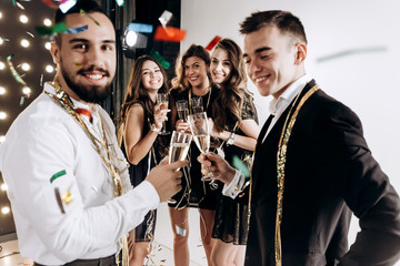 Beautiful young girls and guys dressed in stylish elegant clothes smile  together and clink glasses with champagne confiture around. Party time