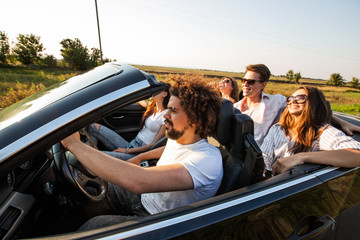 Group of young beautiful girls and guys in sunglasses   smile and ride in a black cabriolet on the road on a sunny day.