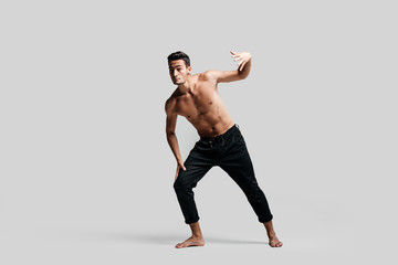 Obraz premium Dark-haired handsome young dancer with bare torso wearing a black pants makes dancing movements on a white background