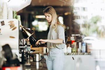A young pretty thin blonde with long hair,dressed in casual outfit,is cooking coffee in a modern coffee shop. Process of making coffee is shown.