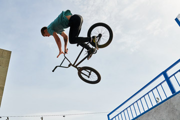 Side view portrait of young man doing bmx stunts jumping up high against sky over ramp in extreme...