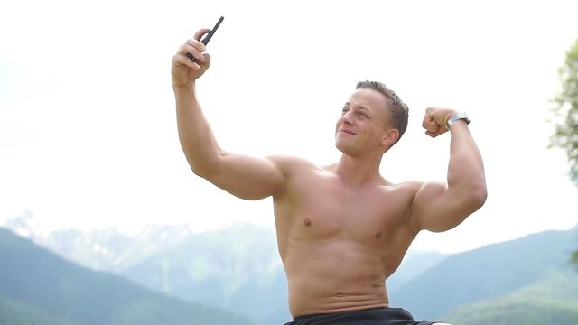 Handsome bodybuilder with perfect muscular body and blonde hair holding smart phone and taking a selfie outdoors over beautiful mountains with green flora on background.