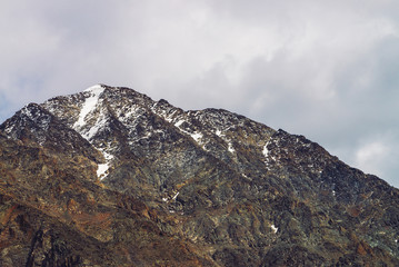 Snowy mountain top in cloudy sky. Rocky ridge under clouds. Overcast weather in highlands. Atmospheric minimalist landscape of majestic nature.