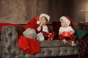 Little cute kids, brother and sister, wearing Santa Claus hats, are sitting on a big easy chair in the living room and are looking at Christmas presents in boxes. 