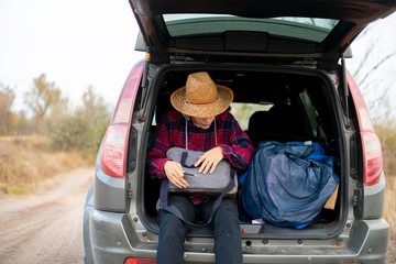 young man in summer hat searching in luggage in the car trunk in road journey f