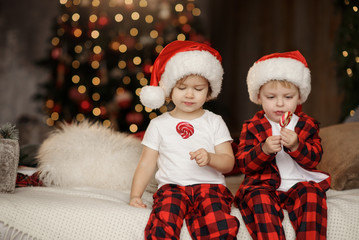 Fototapeta na wymiar Two Little cute children in Santa Claus hat and red pajamas is sitting in the room on the background of the Christmas tree and eating sweet candies on a stick