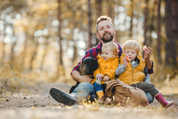 A mature father with toddler children and a dog sitting on the ground in an autumn forest.