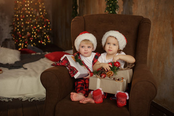 Little cute kids, brother and sister, wearing Santa Claus hats, are sitting on a big easy chair in the living room and are looking at Christmas presents in boxes. 