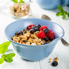 Oat granola with berries and yoghurt in blue bowl on old white wooden background. Healthy breakfast concept.