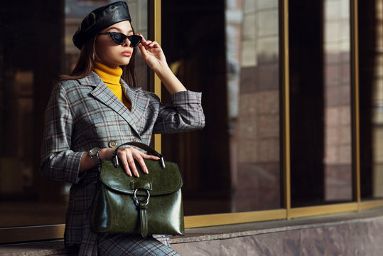 Outdoor fashion portrait of young elegant businesswoman, woman wearing sunglasses, beret, checked suite, blazer, turtleneck, holding green leather bag, posing in street of european city. Copy space