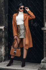 Ganzkörper-Modeporträt im Freien einer eleganten Frau mit Brille, Baskenmütze, Rollkragenpullover, Rock mit Leopardenmuster, kamelfarbenem Mantel, Stiefeletten, Wildlederhandtasche in der Hand, posiert in der Straße der europäischen Stadt © Victoria Fox
