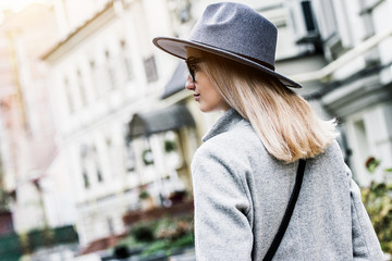Back view of fashionable young woman in coat and hat looking away and walking on the city streets. Attractive and stylish hipster girl in modern urban outfit standing outdoors. City style and fashion
