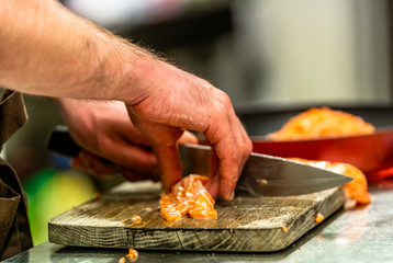 Male Chef Cutting Fresh Salmon on the Wooden Board with Blurred Pan in a Background