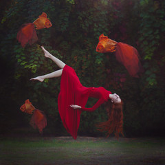 Ein schönes Mädchen mit langen roten Haaren in einem roten Kleid schwebt über dem Boden. Surreales magisches Foto einer Frau mit großem Goldfisch.