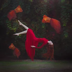 Fotobehang Vrouwen Een mooi meisje met lang rood haar in een rode jurk zweeft boven de grond. Surrealistische magische foto van een vrouw met een grote goudvis.